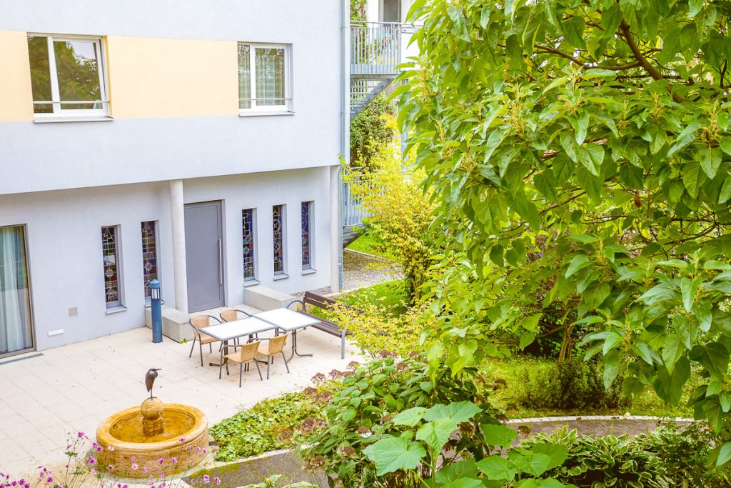 Ein ruhiger Innenhof mit einem kleinen Brunnen, umgeben von üppigem Grün. Das Gebäude hat hellgraue Wände mit hohen, schmalen Fenstern. Auf der Terrasse stehen Tische und Stühle, die einen ruhigen Ort zum Entspannen suggerieren.