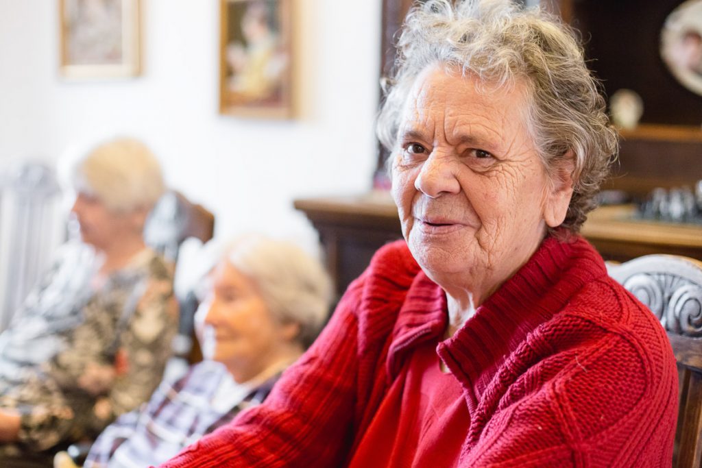 Eine ältere Frau mit lockigem grauem Haar und einem roten Pullover sitzt lächelnd im Haus. Zwei weitere ältere Frauen sind verschwommen im Hintergrund zu sehen und sitzen auf Stühlen. Der Raum wirkt gemütlich, mit gerahmten Kunstwerken an den Wänden.