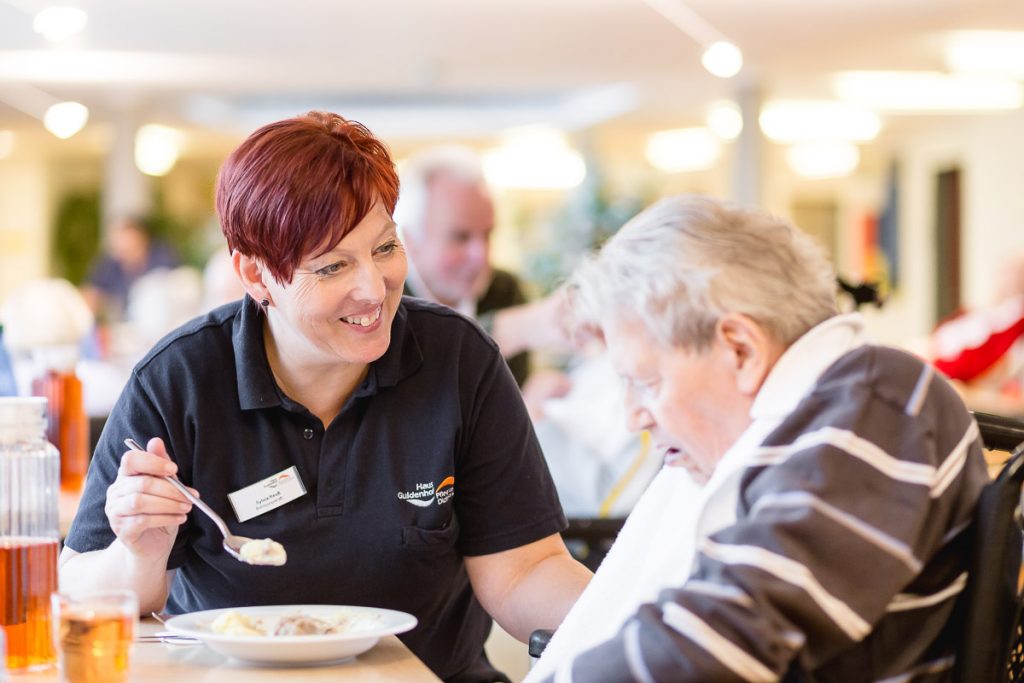 Eine Pflegekraft in einem schwarzen Poloshirt lächelt und füttert eine ältere Person in einem Essbereich. Die ältere Person trägt ein gestreiftes Hemd und ein weißes Lätzchen. Im Hintergrund sind weitere Personen zu sehen.