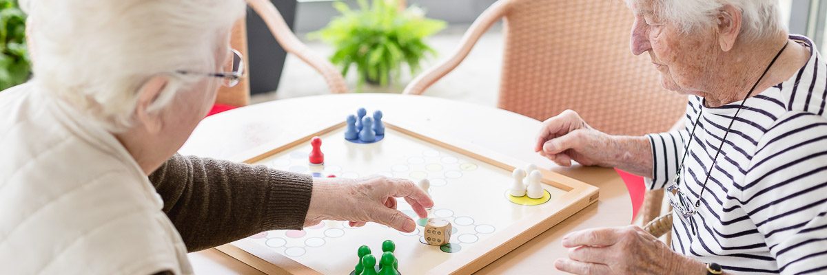 Zwei ältere Personen sitzen an einem runden Tisch und spielen ein Brettspiel mit bunten Spielsteinen. Die Kulisse ist ein heller Raum mit großen Fenstern und viel Grün draußen. Beide sind auf das Spiel konzentriert, einer bewegt einen gelben Spielstein.