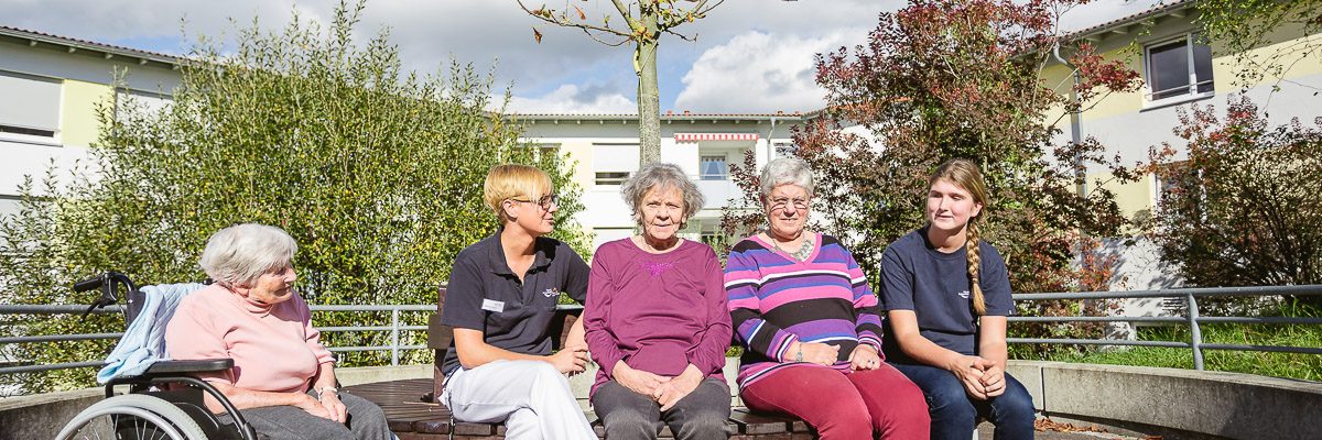 Eine Gruppe von fünf Personen, darunter einer im Rollstuhl, sitzt auf einer geschwungenen Bank im Freien in einem Gartenbereich. Sie lächeln und genießen den sonnigen Tag, umgeben von Bäumen und modernen Gebäuden im Hintergrund.