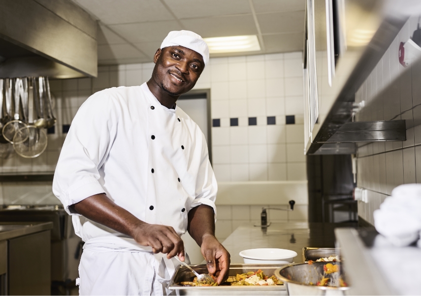 Ein Koch in weißer Uniform und Mütze lächelt, während er in einer professionellen Küche Essen zubereitet. Er hält ein Küchengerät in der Hand und arbeitet mit verschiedenen Gerichten auf einer Metalltheke. Die Küche ist mit Geräten und Werkzeugen aus Edelstahl ausgestattet.