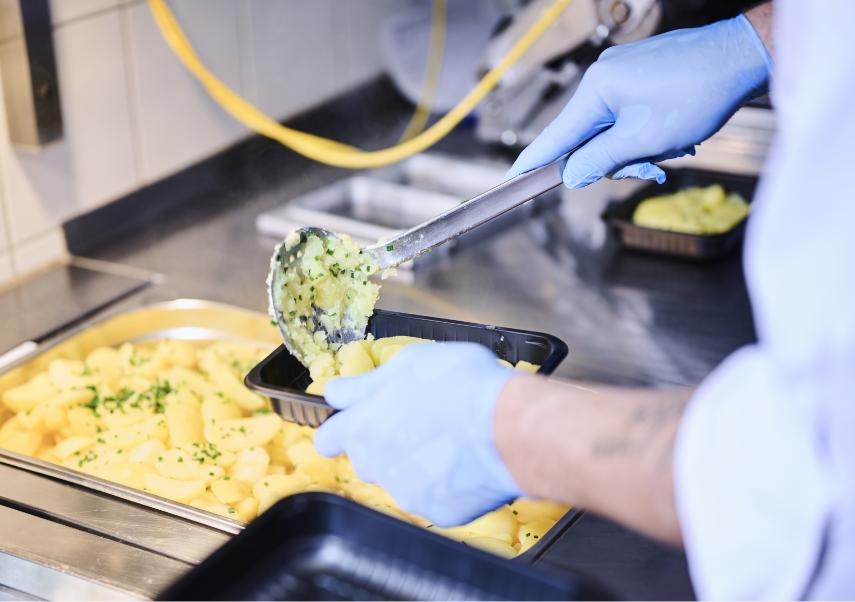 Eine Person mit blauen Handschuhen serviert in einer Küche mit einer Schöpfkelle Nudeln mit grünen Kräutern von einem großen Tablett in einen kleineren Behälter.