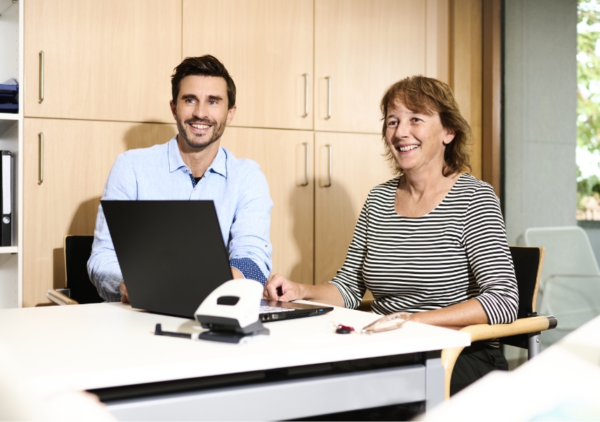 Zwei Personen sitzen an einem Schreibtisch in einem Büro. Ein Mann in einem hellblauen Hemd benutzt einen Laptop und eine Frau in einem gestreiften Hemd lächelt neben ihm. Sie scheinen in einer kollaborativen Umgebung zusammenzuarbeiten.
