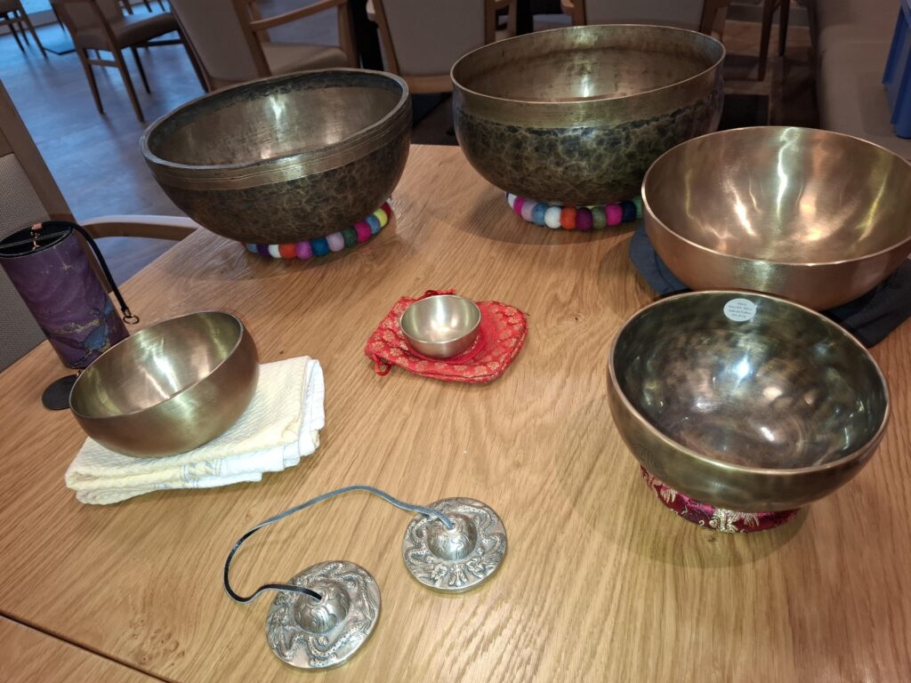Fünf Klangschalen aus Metall in verschiedenen Größen sind auf einem Holztisch angeordnet, einige ruhen auf bunten Kissen. Zwei Metallzimbeln mit Schnüren und ein violetter zylindrischer Gegenstand sind ebenfalls vorhanden. Im Hintergrund stehen Stühle.