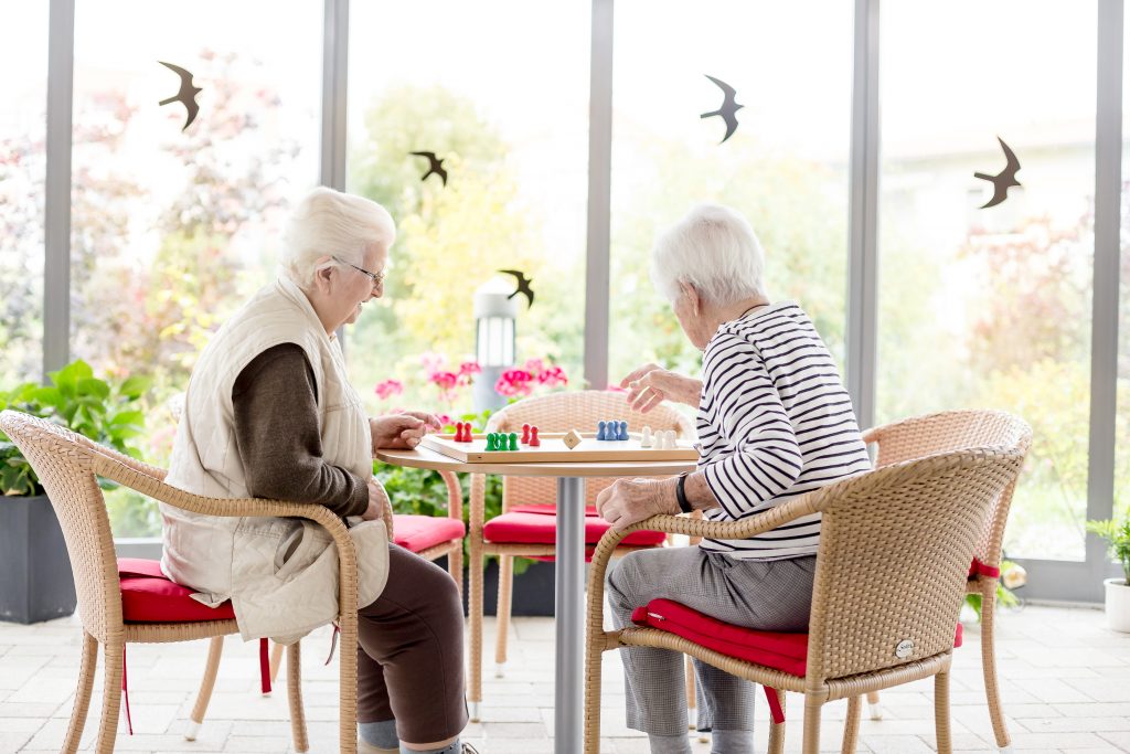 Zwei ältere Frauen sitzen in einem hellen Raum an einem runden Tisch mit einem Brettspiel. Sie sitzen in Korbstühlen mit roten Kissen. Große Fenster geben den Blick auf den Garten frei und an den Wänden hängen Vogelsilhouetten.